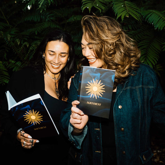 Two Pacific women stand outdoors in front of dark green foliage, each holding a journal with a purple cover featuring a gold sunburst and the title 'ALIGNMENT'. They are smiling as they read what is written in one of the journals.