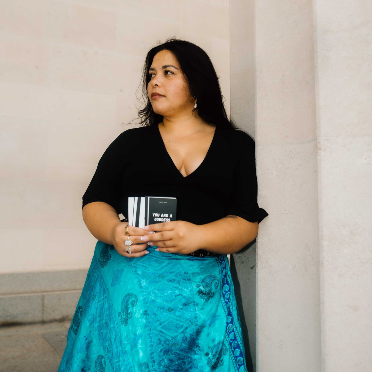 A beautiful Tongan/Maori woman with dark hair stands against a light-colored stone wall, wearing a black short-sleeve V-neck top and a flowing turquoise blue patterned skirt. She is holding a small black and white striped card deck with the first card reading 'YOU ARE A GODDESS'