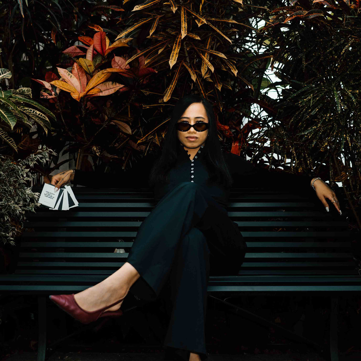 A stunning Samoan woman wearing a black outfit, sunglasses, and burgundy pointed-toe heels sits on a dark green park bench surrounded by lush tropical foliage. She is holding a deck of affirmation cards fanned out with the first one reading 'YOUR OUTPUT IS NOT A REFLECTION OF YOUR WORTH'