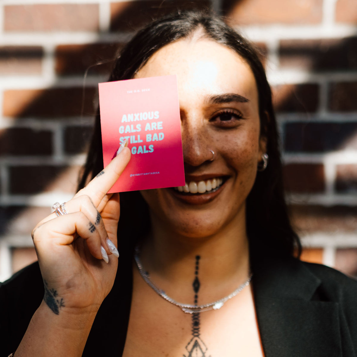 A beautiful Pacific woman smiling in front of a brick wall, holding a pink card with blue text that reads 'ANXIOUS GALS ARE STILL BAD GALS' in front of one eye. She is wearing silver jewellery with long painted nails with a Samoan hand and sternum tattoo.