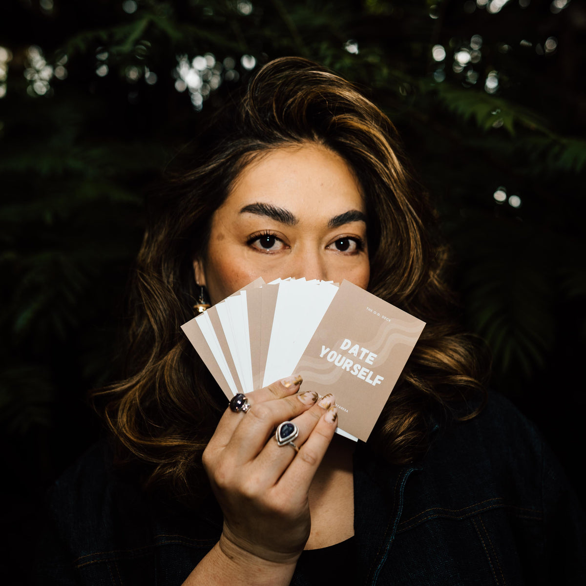 A gorgeous woman holding a fanned-out set of Affirmation cards that read 'DATE YOURSELF' against a dark, blurred outdoor background. She is wearing silver jewellery and has an amazing blowout.