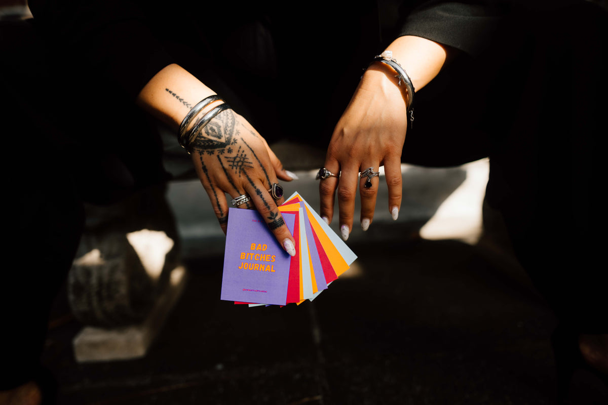 A close-up of tattooed hands holding a fanned-out set of colorful cards or booklets, with one purple card reading 'Bad B!tches Journal' visible. The scene is dark with dramatic lighting, and the hands are adorned with rings and bracelets.