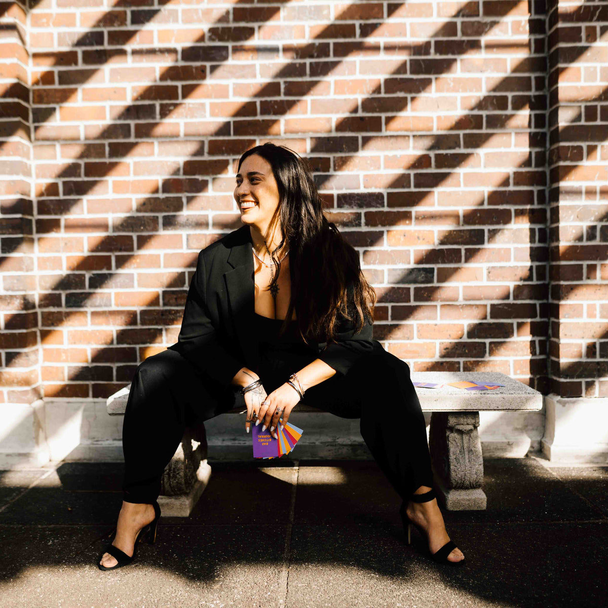A Pacific woman sits on a stone bench against a brick wall, wearing a black blazer, black pants and black heels. She is holding colourful Affirmation Cards which read 'BAD B!TCHES JOURNAL'. holding a small colorful clutch. She has a Samoan hand tattoo and sternum tattoo and is wearing silver rings and bracelets. Strong diagonal shadows from a nearby structure fall across the brick wall and pavement.