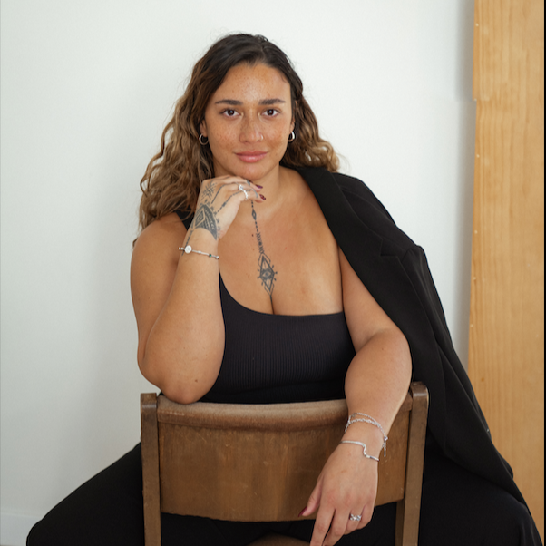 Pacific woman (Dr Britt) sitting on a chair wearing a black suit with tattoos visible on her arms and sternum alongside silver jewellery