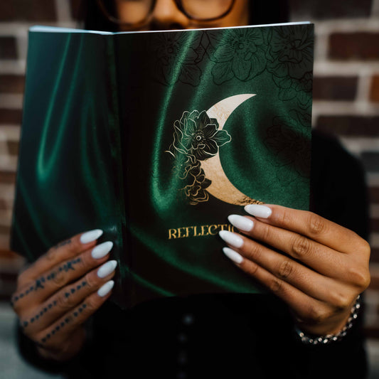 A Pacific woman holding an open journal with a dark green satin-like cover featuring a golf crescent moon and floral illustration, with the word 'REFLECTIONS' visible on the page. The person has long white manicured nails and Samoan tattoos on their hands, siting in front of a brick wall.
